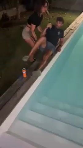 Two people are near a pool. The pool is square or rectangular with concrete steps leading into it. It is lit, and the water is turquoise in color. One person, a man wearing blue shorts, a dark t-shirt, and flip flops, is sitting on the edge of the pool. A person standing behind him, a woman wearing denim shorts and a dark cropped t-shirt, pushes him into the pool. A can of Fanta sits near the pool's edge. The man is holding a beer and drops it as he falls into the pool. The woman laughs. The pool is surrounded by grass and wood decking. The video is shot at night.