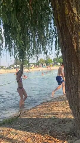 A young girl in a bikini is holding onto the branches of a willow tree, preparing to jump into the water. She is on the sandy shore of a lake, with the water extending out before her. In the background, a beach is visible, populated with people relaxing on towels, swimming, and engaging in water activities. There are also some small cabins or structures on the beach, suggesting a recreational area or campground. The sun appears to be shining, indicating a warm, summer day. The girl’s pose suggests anticipation and excitement for her impending splash into the lake. The scene depicts a leisurely summer day at a popular lakeside destination.