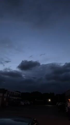 A vertical, low-light video shows a dark, cloudy sky at dusk or dawn over a parking lot. The clouds are thick and dark, suggesting impending rain or a storm. In the lower portion of the frame, a line of parked cars is visible, silhouetted against the dim light. A few buildings are also visible on the left and right sides of the frame. A single street light illuminates a small area in the distance, casting a warm glow. The foreground is partially obscured by the top of a vehicle. The overall mood is somber and atmospheric.