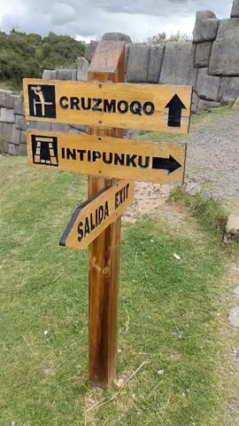 The video shows a stone structure at an archeological site. In the foreground there is a wooden sign post with directions to Ruzmoqo, Intipunku, and the exit. Behind the sign is a large stone wall made of irregularly shaped, gray blocks. A set of wooden stairs leads up and through an opening in the wall. The ground is covered in gravel and patches of grass. The sky is overcast and gray. The camera pans up and slightly to the right to focus on the stone wall.