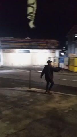 A person wearing a dark hoodie and a baseball cap walks across a street at night. They appear to be holding a thin, dark object in their right hand, possibly a stick or a tool. The street is dimly lit, with buildings and some yellow containers visible in the background. The person is moving from the right side of the frame to the left, and then continues walking away from the camera. There are metal poles on either side of the street.