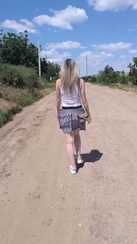 A woman with long blonde hair, wearing a white tank top and a patterned skirt, walks down a dirt road. She has a small bag slung over her shoulder and is wearing white sneakers. The sun is shining brightly, casting a shadow from the woman onto the road. The sky is a clear blue with a few white clouds. Trees and bushes line the sides of the road, and in the distance, there are power lines and some buildings. The scene appears to be in a rural or semi-rural area. The woman's posture suggests a relaxed or leisurely walk. The time of day is clearly daytime due to the bright sunlight and shadows. The weather appears to be warm and sunny. There is no visible text in the media file.