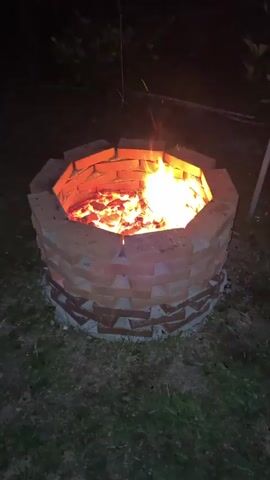 A video shows an octagonal brick fire pit filled with burning embers and a small flame. The fire pit is situated on a patch of grass in a dark, outdoor environment, possibly a backyard, with some foliage visible in the background. The fire pit is constructed with two layers of brick. The lower layer appears to be partially covered with cement, while the upper layer is built higher and forms the octagonal shape. The bricks are laid in a staggered pattern. The fire within the pit emits a warm, orange glow. The lighting suggests it is nighttime or dusk. There are no people or animals visible in the frame. The video is short, lasting only a few seconds, and shows minimal movement of the flames.