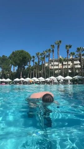 A person is shown underwater in a swimming pool, riding an underwater stationary bicycle. Another person is swimming in the background. There are multiple stationary bicycles in the pool. Palm trees and buildings are visible above the water's surface in the background.