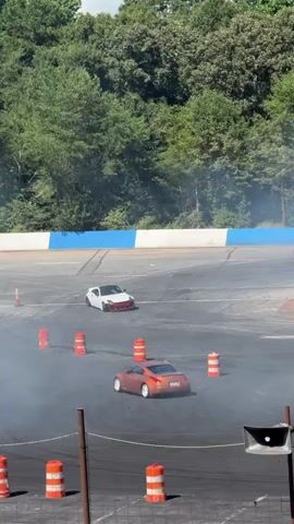 The video shows a professional drifting event. Three cars, one white, one orange, and one silver, are seen drifting around a track. The track is lined with orange traffic cones. In the background, there is a lush green forest and a blue and white barrier. There are also some spectators visible behind the barrier. The cars are kicking up a lot of smoke as they drift, creating a dramatic visual effect. The event appears to be well-attended, and the atmosphere is energetic and exciting. There is a white pickup truck and a yellow front-end loader near the track, suggesting that this might be a professional event with support staff on hand. The cars are all sports cars and appear to be in good condition, with no visible damage. The drifting is skillful and precise, with the cars executing sharp turns and maintaining control. The smoke from the tires is thick and billowing, adding to the visual spectacle. The overall impression is one of a high-octane and thrilling event, with skilled drivers performing impressive maneuvers.