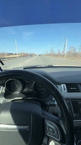 The view from the driver's seat of a Range Rover on a highway shows a sunny, clear blue sky above a straight stretch of road. Other cars can be seen in the distance. The car's dashboard is visible, displaying the speedometer reading around 99 km/h. The steering wheel is prominently featured. Power lines and sparsely wooded areas line the side of the highway.