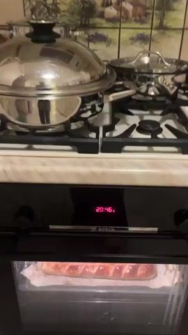 The video opens with a close-up of a stainless steel pot on a gas stovetop. The camera then pans down to an oven with a digital display showing the time 20:46. A hand opens the oven door, revealing a golden-brown, baked pastry on a baking sheet. The pastry is long and rectangular with a diamond-patterned scoring on top, glazed to a high shine. The oven interior is visible, including a circular fan at the back. The camera zooms in slightly on the pastry before the video ends.