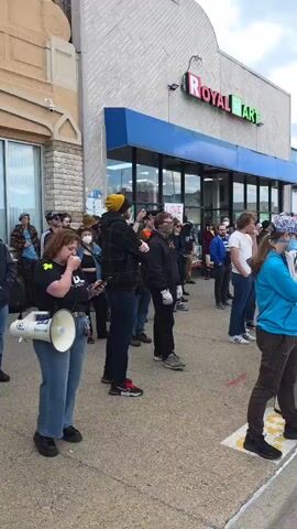 The video shows a crowd of people standing outside a store called "Royal Mart". Several people are holding up their phones, appearing to record the event. A woman with a megaphone is near the front of the crowd. Another man is holding a phone up high, also recording. The camera pans to the left to capture more people in the crowd, some wearing masks and scarves. Some are looking at the camera, others are looking towards the front.