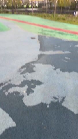 The camera moves slowly along a gray paved walkway next to a green and red path that leads toward several tall buildings in the background. The buildings are lit up with vertical stripes of light. Some puddles of water sit on the walkway. The sky is blue in the distance.
