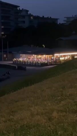 The video begins with a wide shot of a beach scene at dusk. In the foreground, a grassy incline slopes down towards a sandy area. Beyond the sand, a long, elevated deck is visible, filled with people seated at tables. Lights illuminate the deck, creating a warm glow. In the background, there are buildings and dark trees. The scene then transitions to a more distant view of the ocean and beach. The sky shows the colors of a fading sunset, with hues of pink and orange. Waves gently roll onto the shore. Two figures are seen walking on the wet sand near the water's edge, one holding a light. The video continues to pan across the beach, showing the expanse of sand and the ocean.