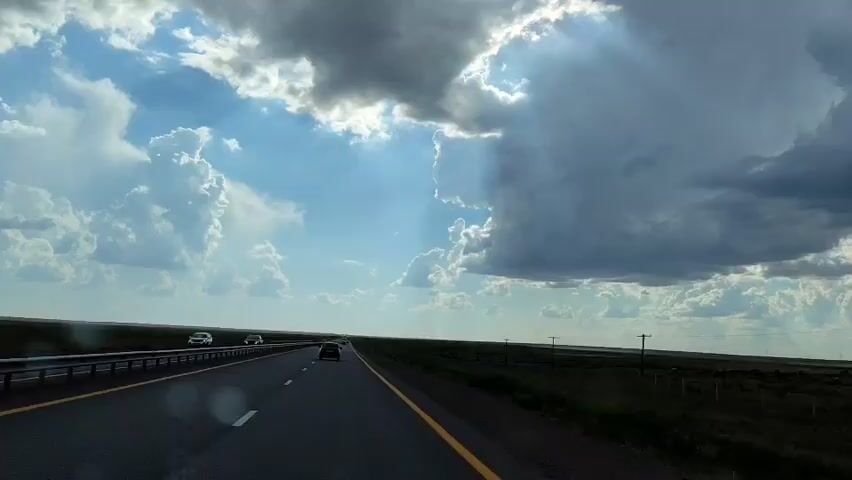 A dashcam view shows a highway with two lanes in each direction. Several cars are visible on the road. The sky is filled with dramatic clouds, with sunlight breaking through in some areas. The landscape on either side of the highway appears to be flat and grassy with some scattered vegetation and utility poles. The overall impression is that of a road trip through an open, potentially rural area under changeable weather conditions.