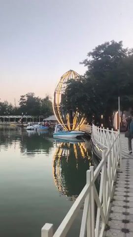 The video pans across a serene lake with several pedal boats scattered on its surface. In the background, there are buildings with lights, trees, and a large, illuminated egg-shaped structure with a Ferris wheel-like design. A white railing runs along the edge of the lake in the foreground, and a pathway is visible behind it. The sky is a soft gradient of pink and blue, suggesting either dawn or dusk. The water is calm, reflecting the lights and the sky.