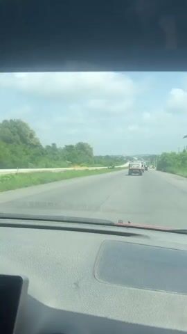 The video is a first-person perspective of a vehicle driving on a paved road. The road has multiple lanes and appears to be in a rural or suburban area, with lush green trees and vegetation on both sides. The sky is partly cloudy. Several other vehicles are visible on the road ahead, including a dark-colored tuk-tuk-style vehicle, a white SUV, and a white van. In the distance, there are some buildings and what appears to be a gas station. The interior of the vehicle from which the video is being recorded is visible in the foreground, including part of the dashboard. The camera is tilted slightly downward, focusing on the road and the vehicles ahead.