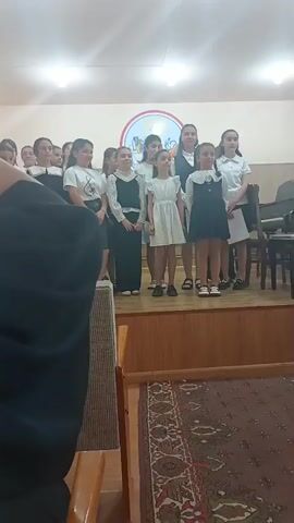 The video shows a group of young girls, likely a choir, standing on a wooden stage in what appears to be a performance hall. They are dressed in uniforms, mostly black and white dresses and skirts, with some wearing white shirts. In the background, a piano is visible to the right of the stage, along with chairs and a wall-mounted circular emblem. The room is lit by ceiling lights, and a patterned rug is spread on the floor in front of the stage. The girls are looking forward, and some of them appear to be singing or about to sing. The perspective is from someone in the audience, looking up at the stage.