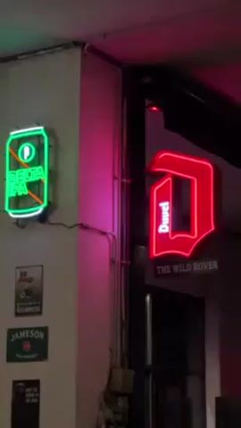 Here's a description of the video:

The video shows the exterior of a bar or pub, likely at night or during the evening, featuring various neon signs.

On the left, mounted on a light gray wall, there is a green neon sign shaped like a beer can displaying "DELTA IPA". Below it, there are several smaller, dark-colored plaques with beer and liquor brand names, including Guinness and Jameson.

To the right, an entrance to the bar is visible. Above the entrance, there is a red neon sign in the shape of a stylized letter "D" with the word "Duvel" inside. Underneath the "Duvel" sign is a plaque that reads "THE WILD ROVER". 

A pink-purple light is cast on the wall between the two neon signs. The overall aesthetic is dark, with the neon signs providing the main sources of illumination. There are also some utility pipes and electrical boxes visible on the wall.