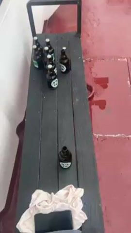 The video shows a scene on the deck of a boat. There are several bottles of beer on a bench, and a large spool of blue rope sits nearby. A person wearing a black hooded sweatshirt is grilling sausages on a barbecue near the edge of the boat, overlooking the sea.