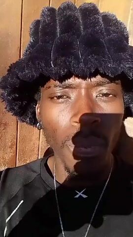 Here is a concise description of the video:

The video shows a dark-skinned person with a mustache and a goatee wearing a black T-shirt, a silver chain, and a large, fuzzy black hat. The person is standing outside with a wooden door to their left and a brick wall behind them. The person looks at the camera, smiles widely, and then looks down. There is music playing in the background.