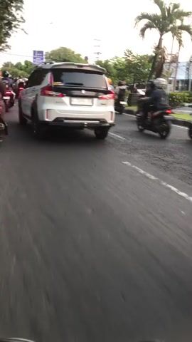 The video shows a street scene with several motorcycles and a car. The perspective appears to be from inside a moving vehicle, likely another car or motorcycle. On the left, a white SUV is visible from the rear. In the center and right, several motorcycles are moving. One motorcycle in the center has a rider wearing a blue jacket and a white helmet. Another motorcycle to its right has two people on it, both wearing helmets and dark clothing. The motorcycles are on a paved road, and there are trees and buildings in the background. The sky is visible and appears to be overcast or during twilight. The overall impression is a busy street with traffic.