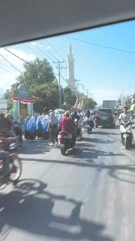 A video shows a street scene with people and motorcycles. A mosque with a green dome and a minaret is visible in the background. Several individuals, some wearing white and others in green, are gathered on the side of the road. Many motorcycles are present, with some people riding them and others parked. The sky is clear and blue. The video appears to be filmed from a moving vehicle.
