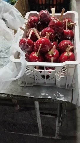 The video shows a white plastic crate filled with at least a dozen shiny, red candy apples. Each apple is on a stick and individually wrapped in clear plastic. The crate sits on a worn, wooden table with a metal frame. The background is slightly out of focus, showing other crates with produce.