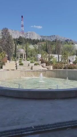 The video shows an outdoor park with a tiered fountain in the foreground. The fountain has a shallow, circular pool with small water jets spraying upwards. Behind the fountain, there are terraced levels with greenery, flowers, and a gazebo. In the distance, a mountain range is visible against a clear blue sky. A tall red and white communication tower stands on one of the mountains. The camera slowly pans to the left, revealing more of the park and the surrounding landscape. The ground near the camera is paved with grey concrete.