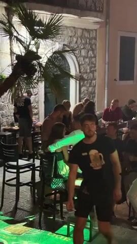 The video shows an outdoor gathering at night, likely a bar or restaurant, with stone buildings and foliage in the background. Several people are seated at tables and standing around, some appear to be engaged in conversation. A man in a black t-shirt with a beer graphic on it is walking towards the camera. Another man in a white t-shirt is partially visible in the foreground, facing away from the camera. Green lights are illuminating some of the seating areas. The overall atmosphere seems relaxed and social.