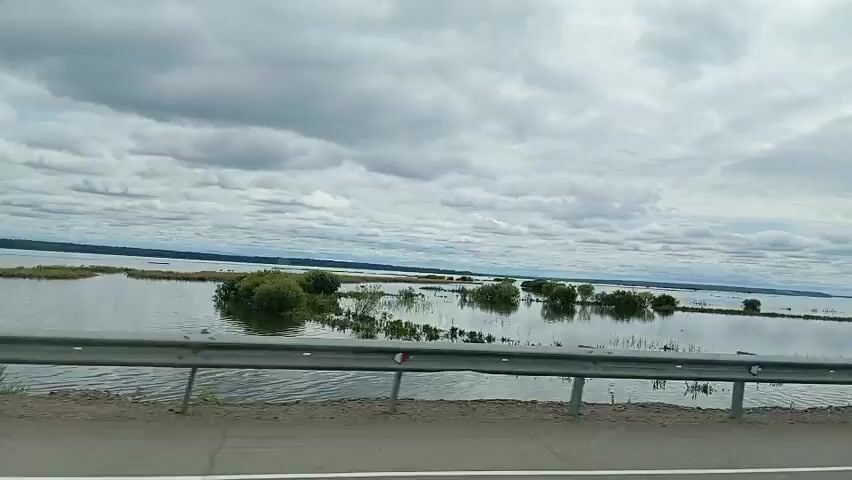 The video shows a view from a moving vehicle, likely a car, traveling along a road. In the foreground, a metal guardrail runs horizontally across the bottom of the frame, separating the road from the landscape beyond. The landscape is dominated by a vast body of water, appearing to be a lake or a flooded area, under a cloudy sky. Several small islands or patches of land covered with vegetation are visible scattered across the water. In the distance, a line of trees marks the horizon. The water reflects the overcast sky, creating a muted and somewhat somber atmosphere. The vehicle continues to move, indicated by the slight blur in the background and the changing perspective. Partway through the video, another vehicle, a large truck with a silver cargo container, briefly comes into view, passing by the camera. The scene suggests a journey through a waterlogged or flood-prone region.