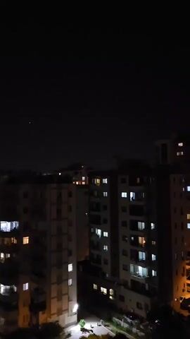 This is a video of a city skyline at night. The sky is dark and there are several buildings with lights on in the windows. The buildings are of various heights and appear to be apartment buildings. There are also some trees and streetlights visible in the lower part of the frame. The camera is panning slowly from left to right, revealing more of the city.