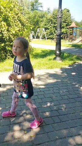 A young girl with her hair in a ponytail, wearing a dark t-shirt with a floral design, light pink leggings, and pink Crocs, stands on a paved walkway. She is looking to her left, with her right hand near her chest. A dark stroller is partially visible in the lower left corner of the frame. Behind her, there is a lush green hedge and trees. Further back, a white swing set is visible. To the right, a black lamppost stands near the walkway. The ground is paved with rectangular stones, and shadows are cast on the ground. The girl then turns her head and looks to her right. A small orange and black bicycle is parked to her right. She continues to walk, looking to her right. In the background, there are more trees and green grass. She then turns her head to her left and looks forward. The video pans slightly to the right, showing more trees and a park bench in the distance. The girl is now holding a red object in her right hand. She continues to walk and then turns her head to the left. A person is sitting in a chair in the foreground, with their legs visible. The girl looks to her left, and the camera pans to the left. She is now holding a black can in her right hand. The background shows trees and a park setting.