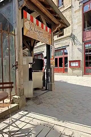 A person walks past an ice cream kiosk labeled "le kiosque à glaces" in a cobblestone courtyard. Behind the kiosk, a building with red trim and large windows can be seen. Another person walks towards the building, followed by two children. The sun casts shadows on the ground.