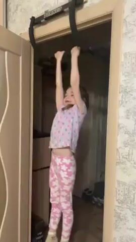 A young girl, wearing a light-colored shirt with small patterns, pink pants with white flowers, and light-green socks, hangs from a pull-up bar attached to a doorway. The pull-up bar is black with the word "TORNEO" on it. She does one pull-up, lifting her body so her chin is above the bar. The doorway leads into a room with shelves visible inside. The walls are covered in wallpaper with a floral pattern.