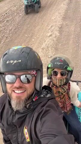 The media shows a selfie-style shot of two individuals riding a quad bike on a dirt road, likely during an excursion in Lalla Takarkoust, Morocco. In the foreground, a man with a beard and reflective sunglasses is smiling broadly at the camera. He wears a black helmet with "VEGA" branding and a black jacket, with a red and white bandana visible around his neck. Behind him, a woman wearing sunglasses, a green "VEGA" helmet, and a colorful paisley scarf covering her lower face waves her right hand. She is dressed in a light-colored top and blue jeans.

The background reveals a wide, unpaved dirt road winding through a dry, rural landscape with sparse vegetation and distant hills under an overcast sky. Several other quad bikes with riders are visible further down the road, indicating a group activity; one in the middle distance is green, and another further back is orange. The side of the ATV in the foreground shows partially visible text "KING". The overall atmosphere suggests adventure and enjoyment in an arid, natural setting.