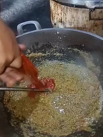 A hand is adding a dollop of red paste to a pan of golden-brown food. The pan is on a stove. A man wearing a black t-shirt is visible in the foreground, partially blurred. The environment appears to be a kitchen.