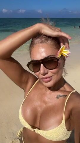 A woman with blonde hair is posing for a selfie on a beach. She is wearing a yellow bikini and sunglasses, and has a flower tucked behind her ear. The ocean and sand are visible in the background. She is touching her hair with her right hand.