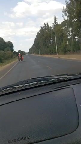 The video shows a view from inside a car, looking out at a paved road under a partly cloudy sky. A motorcycle with a driver and passenger is in the distance ahead on the same road, traveling in the same direction as the car. The motorcycle has a red bag or package attached to the back. The road is bordered by trees on the right and some vegetation on the left. Utility poles are visible along the right side of the road. The dashboard of the car is visible at the bottom of the frame.