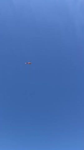 A distant orange object with blades appears to be flying in a clear blue sky. The object is small in the frame, and its details are unclear due to the distance. The sky is a uniform, bright blue, indicating a sunny day with no clouds visible. The footage is static, with the object maintaining a consistent position in the sky, suggesting it is either stationary or moving very slowly relative to the camera.