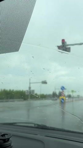 The video shows a view through a car windshield on a rainy day. Raindrops are visible on the glass. The road ahead is wet and reflective. Traffic lights are visible, some showing red. Several cars and a truck are on the road. Road signs, including a pedestrian crossing sign, are also present. Trees and buildings are visible in the background, blurred due to distance and rain. The sky is overcast and gray. The camera appears to be positioned from the driver's seat.