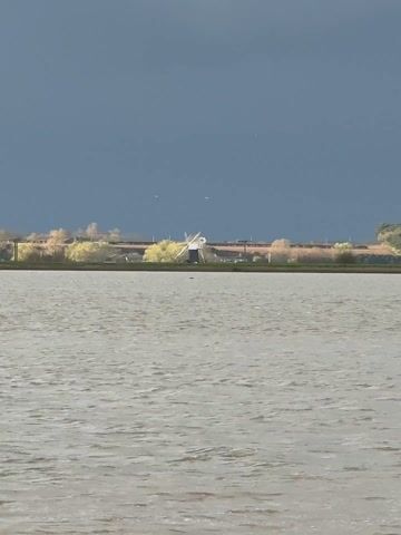 The video displays a wide, static shot of a tranquil landscape. In the foreground, a vast body of water, likely a lake or estuary, occupies the lower half of the frame. The water's surface is lightly ruffled by wind, appearing a muted, light gray color. Above the water, a strip of land runs horizontally across the middle of the scene. On this land, a prominent white windmill with visible blades stands slightly to the left of the center. Behind the windmill and spanning the land strip, there's a linear feature that could be a road or railway track. Scattered along this line are trees or bushes, some of which appear to have yellowish or light-colored foliage. The sky above is a deep, uniform dark gray, suggesting heavy cloud cover or an approaching storm, contrasting sharply with the lighter tones of the water and land. Several small, white birds can be seen flying in the distant sky against the dark backdrop. The scene evokes a sense of quiet solitude and natural beauty.