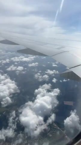 A view from an airplane window shows the wing and clouds below. The ground appears to be a mix of green foliage and some visible buildings and roads. The clouds are scattered, allowing patches of the landscape to be seen. The sky is blue with some wispy clouds.