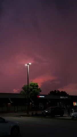 The video shows a dark parking lot with a motorcycle rider navigating through the space. The sky above is a deep purple with hints of red and orange, suggesting a sunset or twilight. A few streetlights are visible, casting a dim glow on the scene. Trees and buildings line the background, partially obscured by darkness. The motorcycle is moving, its taillight illuminated, and the rider appears to be alone. The overall mood is dim and atmospheric, capturing a fleeting moment at the end of the day.