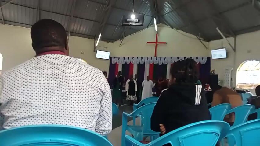 The video shows a church interior with a group of people gathered at the front. A red cross is visible on the back wall, above a backdrop of colorful fabric. Some individuals are standing in a line, while others are seated in blue chairs. The lighting is artificial, and the camera is handheld, resulting in a slightly shaky image. The overall atmosphere appears to be that of a religious service or gathering.