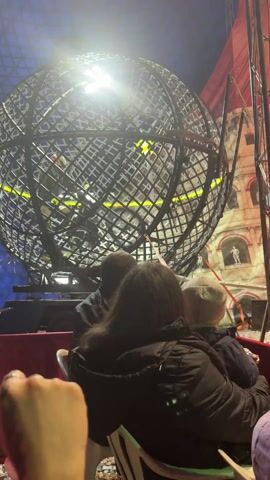 From the perspective of an audience member, a circus attraction is underway. Three people, probably a parent and two children, are seated in front of the camera, watching a large metal globe. Inside the globe, a motorcycle rider is performing stunts. The globe is brightly lit, and behind it, there is a backdrop of a building facade.  The sound of the motorcycle engine is audible. The overall environment suggests a circus or similar entertainment venue.