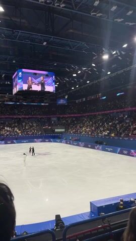 The video shows a figure skating competition in a packed arena. A pair of skaters are performing on the ice. A large screen above the ice shows a close up of the skaters. The stands are filled with spectators. A photographer stands near the edge of the ice. The backdrop has text that reads "Milano Cortina 2026" with the Olympic rings. The skaters wear elegant costumes and glide across the ice in sync.