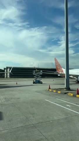 The video shows two airport workers in a raised scissor lift performing maintenance on the wing of an airplane. The scissor lift is a blue Genie GS-2669 RT 4x4 model, and the workers are wearing high-visibility clothing. The airplane has a red and brown patterned tail. The backdrop includes a large, gray building and a blue sky with scattered clouds. Orange cones are placed on the ground in the foreground. The camera appears to be recording from a moving vehicle.