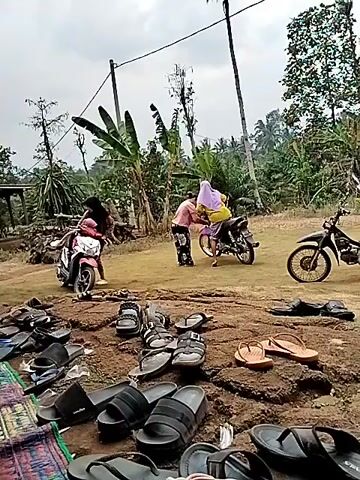 In an outdoor, rural setting with dirt ground and lush vegetation in the background, several people and motorcycles are visible. In the foreground, a large collection of various sandals and flip-flops are scattered on the ground. In the mid-ground, two individuals are interacting near a motorcycle. One person, wearing a pink shirt and black pants, appears to be assisting another person, who is seated on the motorcycle and wearing a yellow shirt and a light purple head covering. A second motorcycle is parked to the right. Further to the left, another person on a motorcycle is visible. The environment suggests a casual gathering or event.
