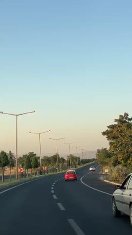 The video shows a view from a moving vehicle on a two-lane road. The road curves to the left. Several cars are visible on the road. The sky is clear and blue. Streetlights line the left side of the road, and trees and bushes are present on both sides. There are also some buildings and structures visible in the distance. The time of day appears to be morning or late afternoon.