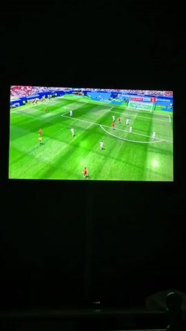 A television screen displays a soccer match. Two teams are playing. One team wears white uniforms and the other wears red and yellow uniforms. The match is being played on a green field with white lines. The scoreboard indicates it is a FIFA World Cup match. The screen then zooms in to show two players, one in a white uniform and the other in a red and yellow uniform, looking at each other.