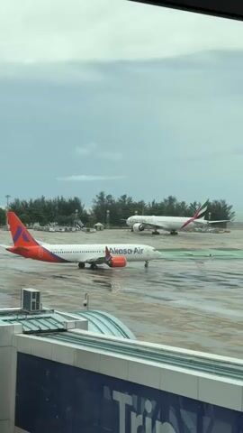 This video captures an outdoor view of an airport tarmac on a cloudy day. Two airplanes are visible on the wet asphalt. In the foreground, a white airplane with orange and purple markings, identified as "Akasa Air" by its livery, is positioned on the tarmac. Further in the background, a larger white airplane with red and green tail fins is also present. The ground appears wet, suggesting recent rain. The scene is framed by the window of an airport terminal, with a blue and white structure visible in the lower portion of the frame. Trees and some buildings are discernible in the distance behind the airplanes. The overall atmosphere is overcast and somber.