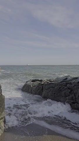 A vertical, medium shot captures waves crashing against dark, rocky shorelines under a bright, overcast sky. A distant cargo ship is visible on the horizon. The water is choppy, with white foam and spray. The shoreline is composed of dark, textured rocks and a patch of sand or gravel in the foreground. The scene is peaceful and natural.