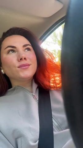A woman with red hair is sitting in a car, wearing a grey zip-up sweatshirt and a seatbelt. She glances around, smiles, and then raises a hand to adjust the car's sun visor. Outside the window, trees flash by, suggesting the car is in motion on a sunny day. The sound of upbeat Russian music fills the background.